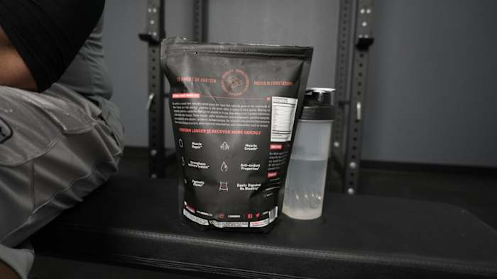 The backside of a bag of XWerks Grow whey protein powder and a shaker bottle with water on a weight bench in a gym